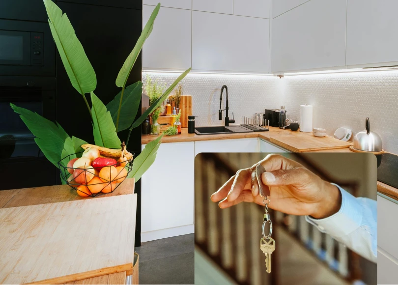 Modern minimalist kitchen with white cabinets, black appliances, and light wood countertops featuring a black sink with modern black gooseneck faucet and plumbing fixtures. A hand holding house keys is visible in the bottom right corner, suggesting new home ownership. The kitchen includes under-cabinet LED lighting, white hexagonal tile backsplash, and various kitchen items including a coffee machine, fruit basket, and green plants.