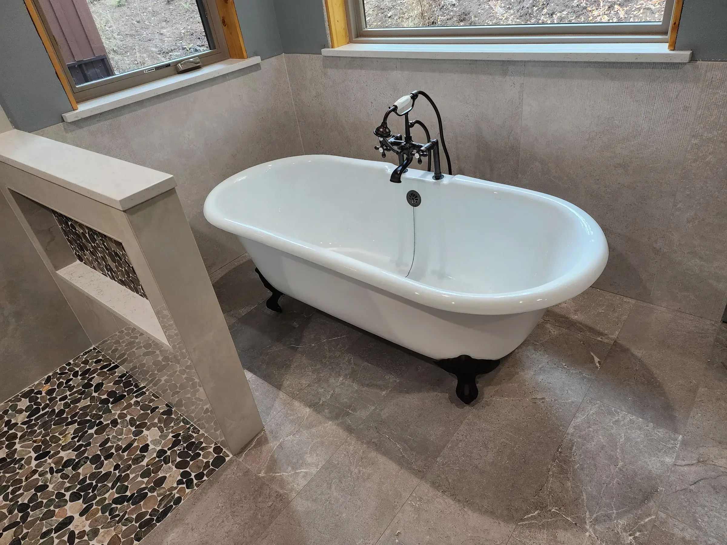 A finished bathroom with a freestanding clawfoot tub, large gray tile and a pebble shower floor