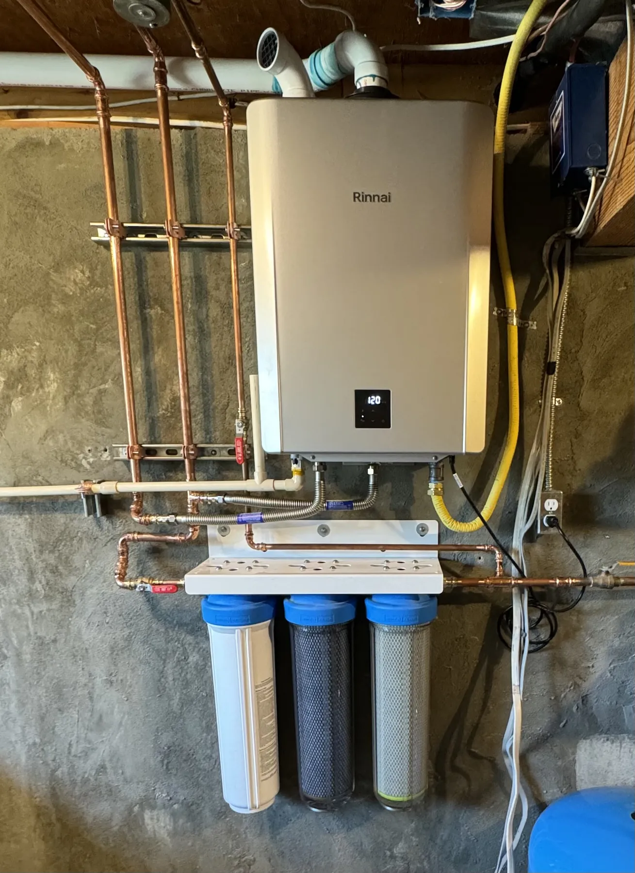 A Rinnai tankless water heater mounted on a copper manifold above three water filter housings