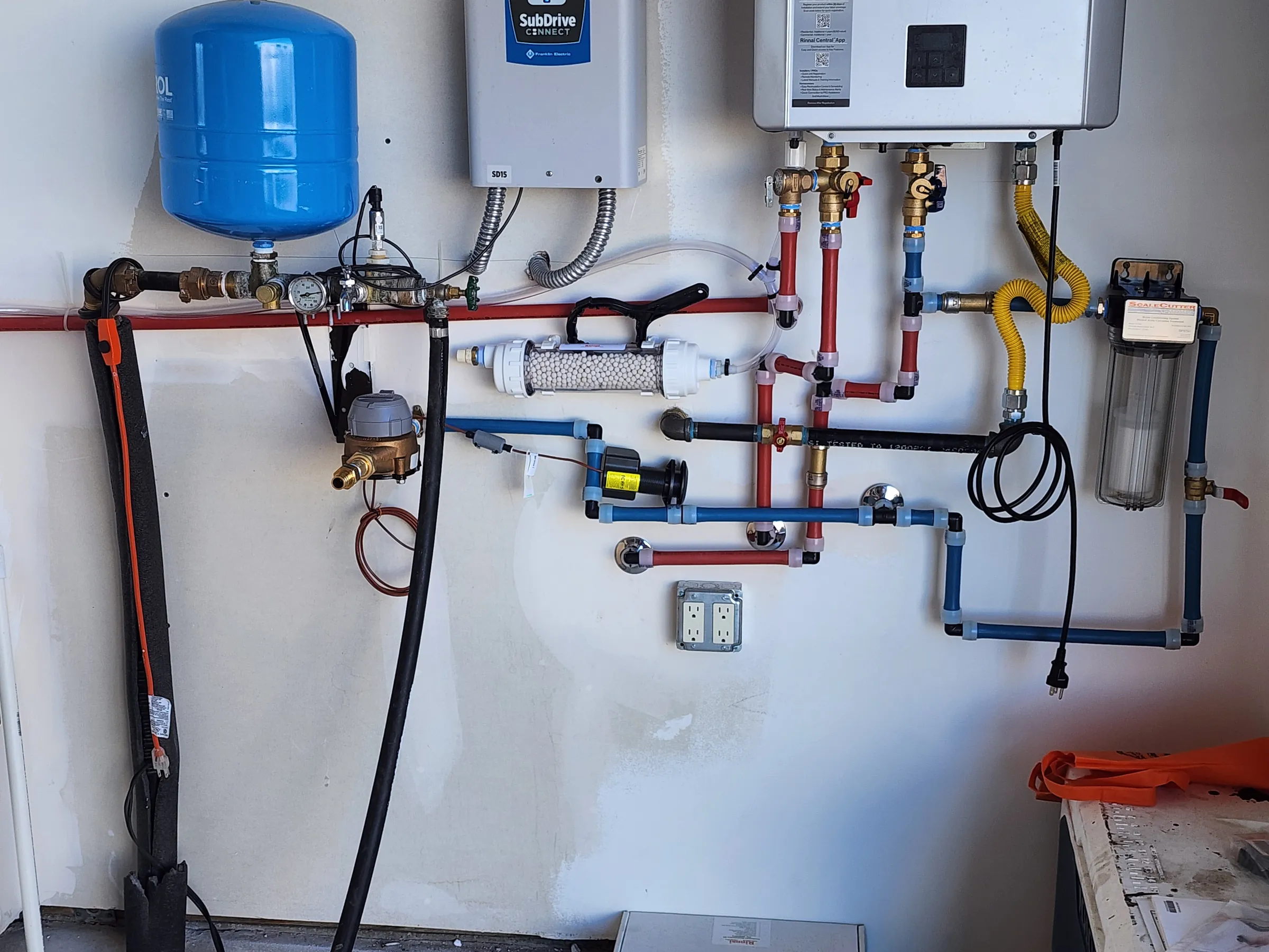A tankless water heater, blue expansion tank and filter housings plumbed on a white wall in red, blue and copper lines